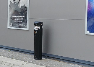 Ashguard™ SG Cigarette Bin in black in front of building with posters for events on building wall