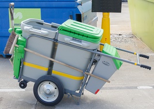 Double Space-Liner™ Orderly Barrow in grey with green lids and detailing with litter picker and broom attached and operator pushing it on footpath