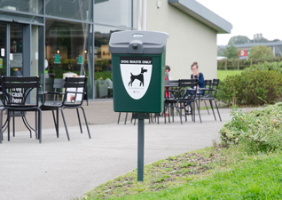 Fido™ 25 Dog Waste Bin in deep green with post mounted fixings onto grass area next to seated area outside motorway services