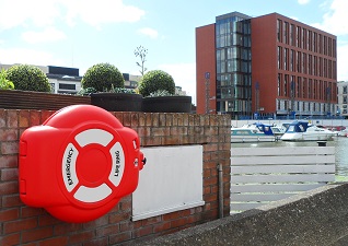 Guardian™ Lifebuoy Cabinet in red with white stickers on, fixed onto brick overlooking marina and modern building