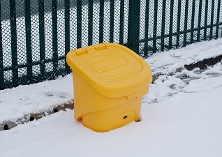 Nestor™ 90 Grit Salt Bin in yellow next to green railing on snowy ground