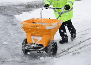 Turbocast™ 300 Manual Salt Spreader in yellow being pushed on snowy ground by operator in high visibility jacket